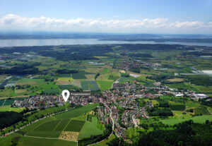 Idyllische Landschaft eines Dorfes mit grünen Hängen, Feldern und einem See unter einer wolkenverhangenen blauen Wolkendecke.